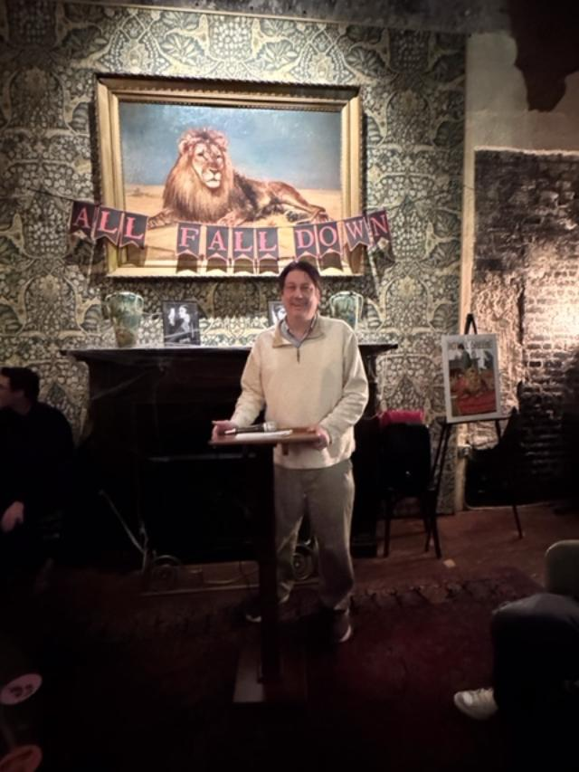 Bruce Buchanan reading at the podium of the Hotel Greene, with a painting of a lion over his shoulder