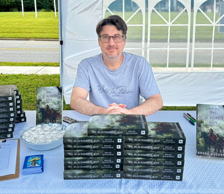 Author Bruce Buchanan sits behind an outdoor table with copies of his book, The Blacksmith's Boy